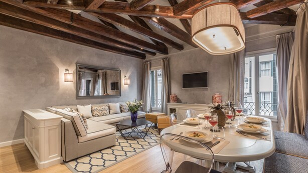 Una sala moderna con sofá, mesa de comedor y sillas. Tiene un ventanal con cortinas, un televisor de pantalla plana y una araña.