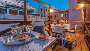 Terraza al aire libre con mesa lista para cenar, exhibiendo una bandeja con vasos y postres, con vista a un paisaje urbano al atardecer.