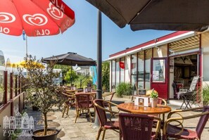 An outdoor cafe area with wicker chairs and tables, umbrellas, and a building with a red awning in the background.