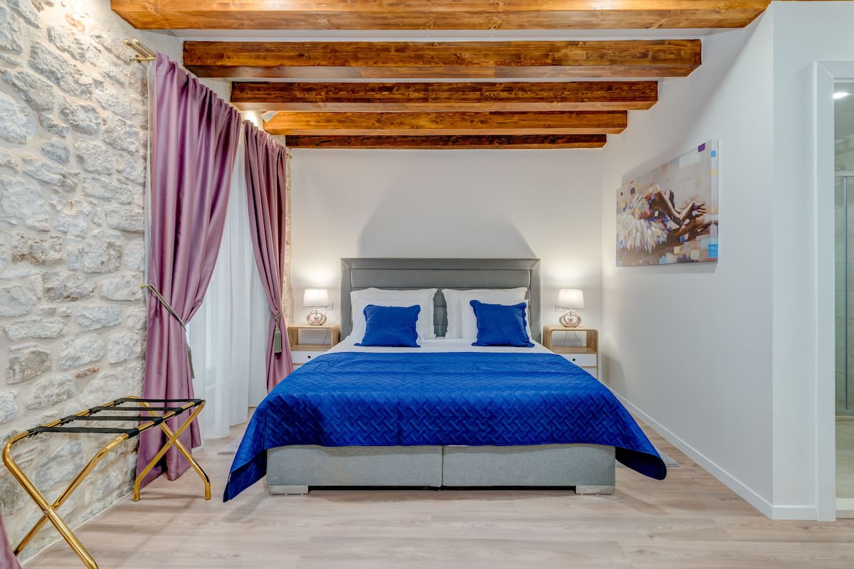 A bedroom with a stone wall, wooden ceiling beams, a bed with blue bedding, a nightstand with a lamp, and a painting on the wall.