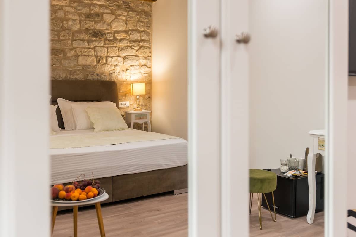 A bedroom with a stone wall, wooden beams, a bed with pillows, a bedside table with a lamp, a small table with fruit, and a chair.