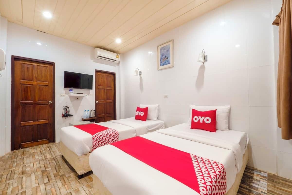 A hotel room with two beds, each with a red and white bedspread and pillows with the brand name "OYO". There is a wooden door, a television mounted on the wall, and an air conditioning unit above it.