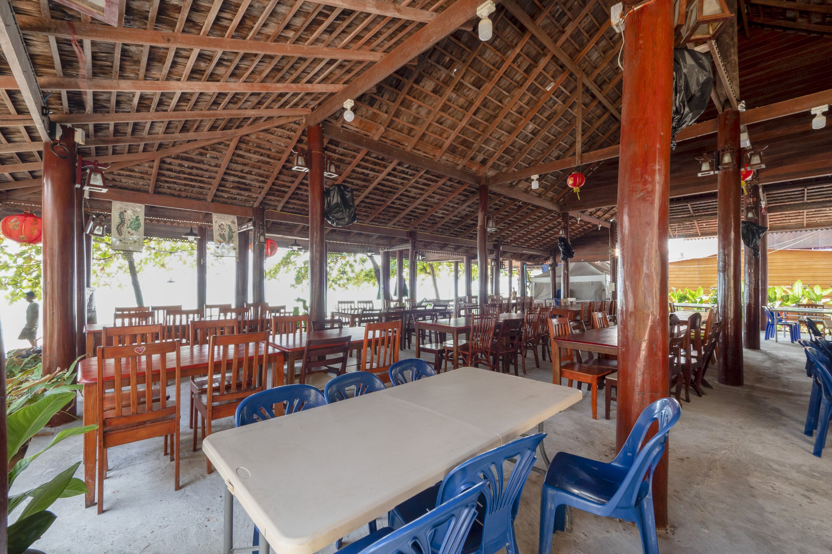An indoor dining area with wooden tables and chairs, a tiled floor, and a thatched roof structure.