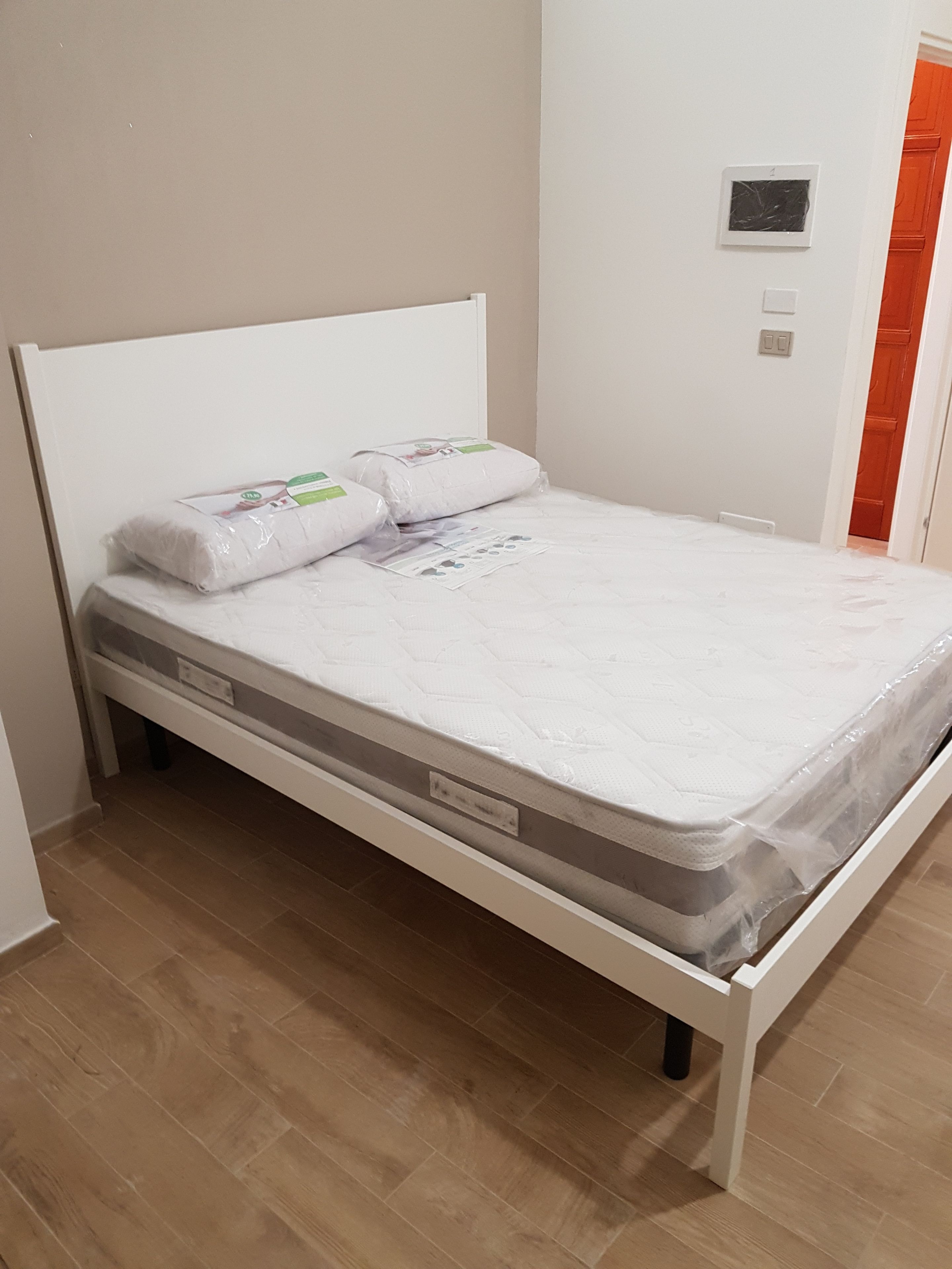 A single-bed room with a white headboard, a mattress with a protective cover, and a wooden floor.