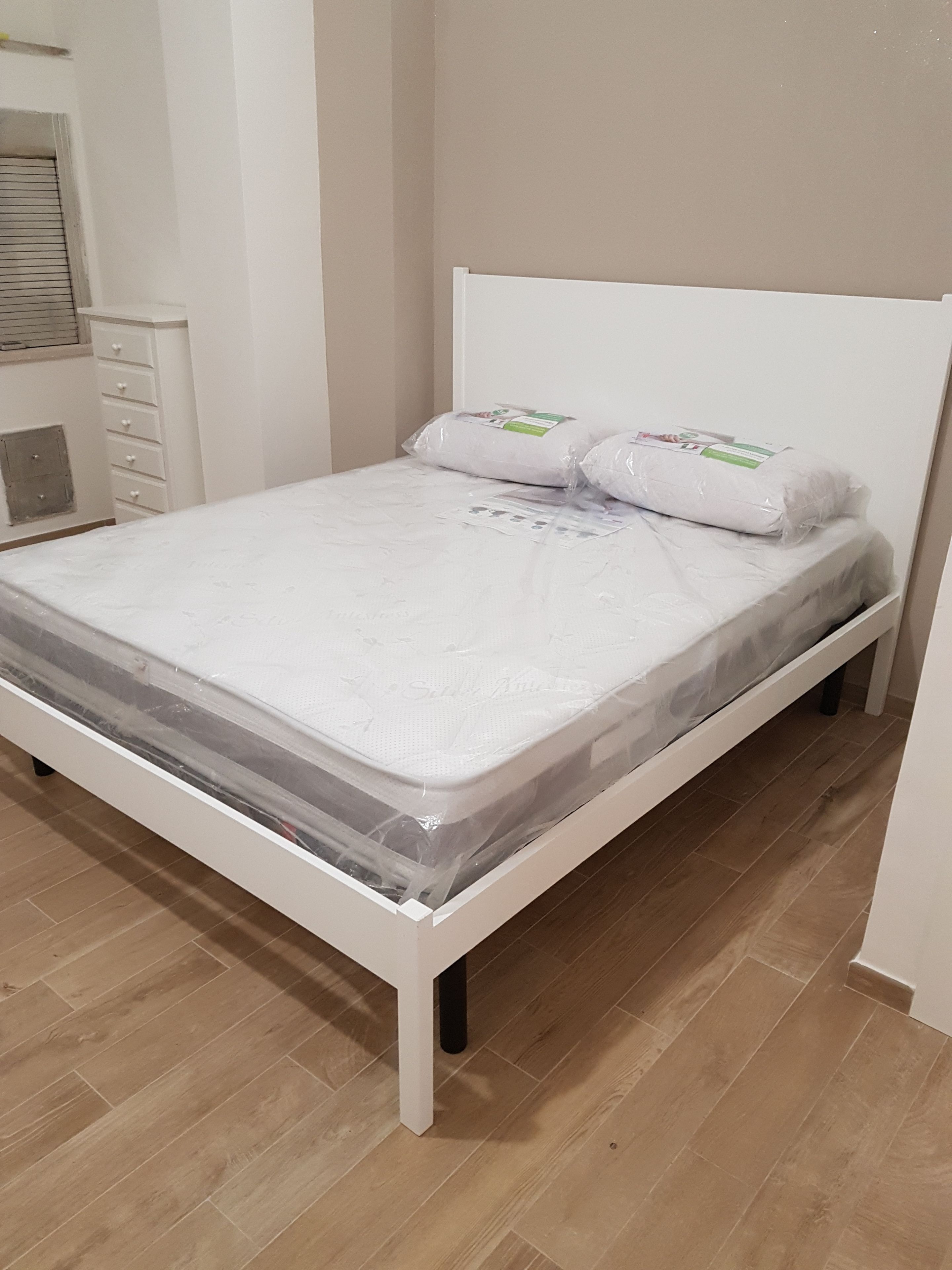 A white bed with a mattress cover, two pillows, and a headboard in a room with wooden flooring and a white wall.