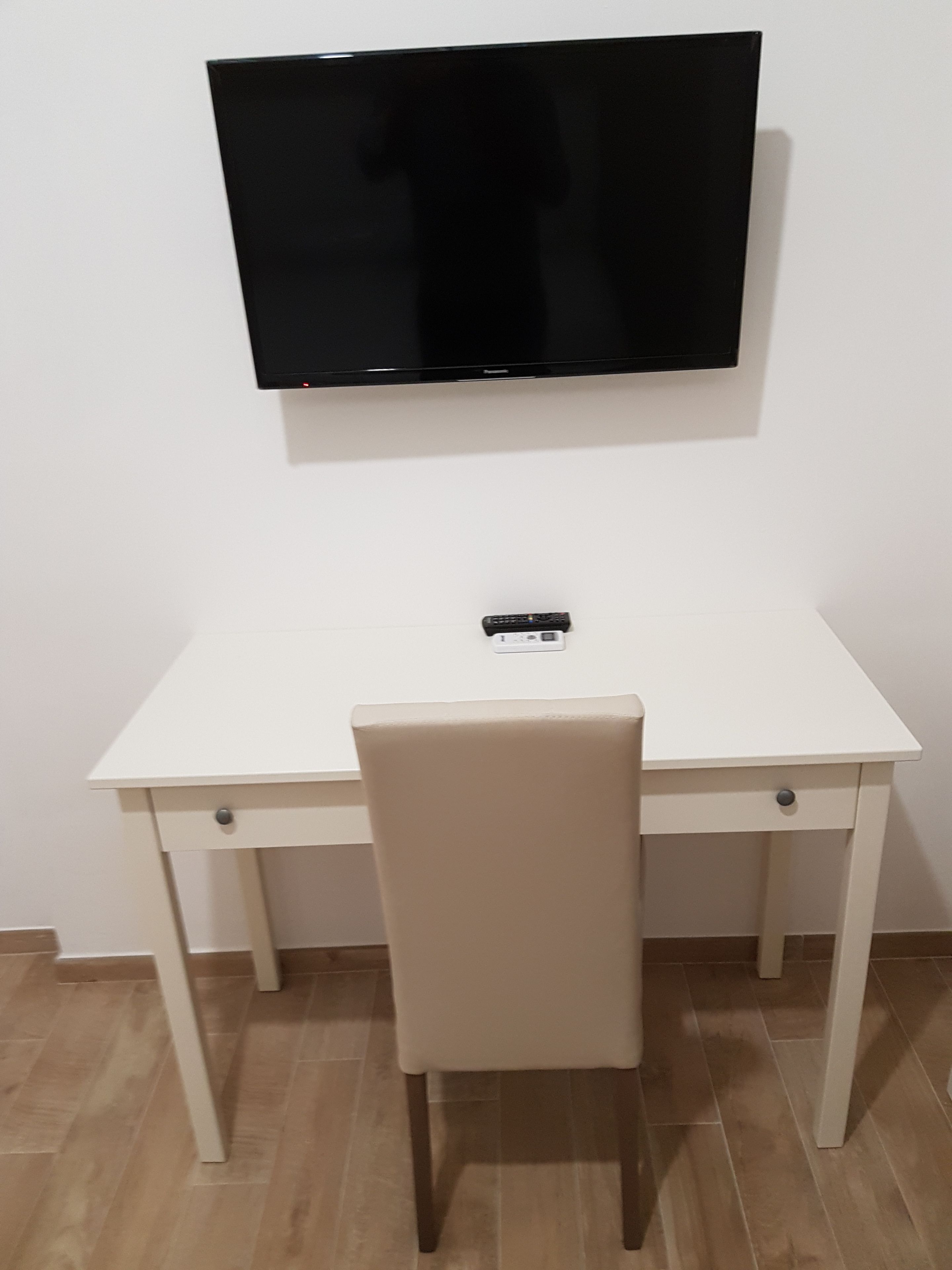 Una scrivania bianca con una sedia beige, una TV a schermo piatto montata sopra e una presa elettrica sulla parete.