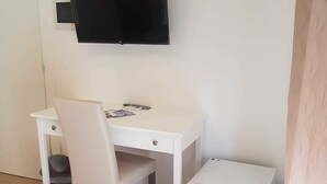 Una stanza piccola e pulita con una scrivania bianca, una sedia, una televisione a parete e un frigorifero bianco.