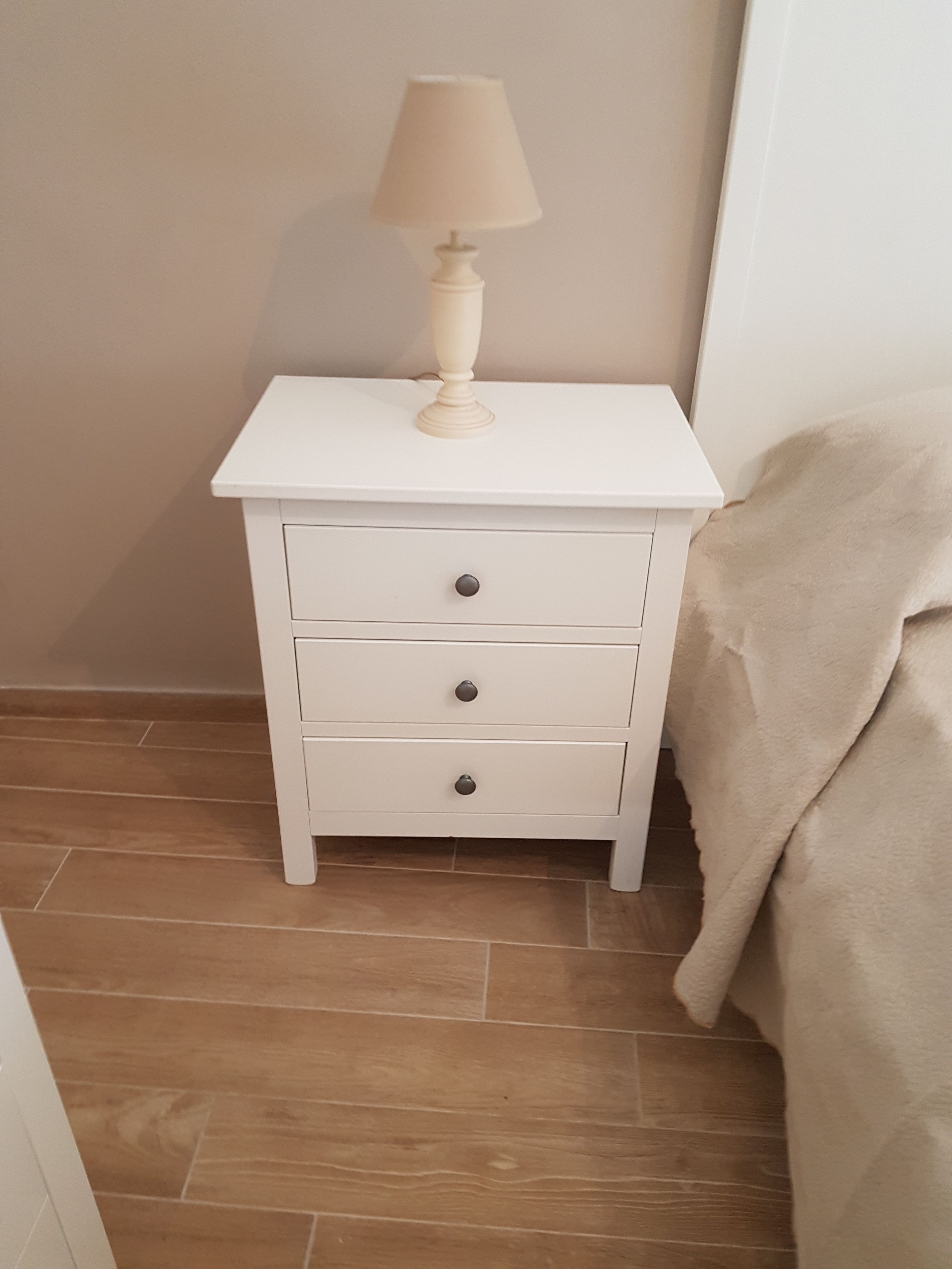 Una mesita de noche blanca con una lámpara, tres cajones y una manta color beige sobre una cama.