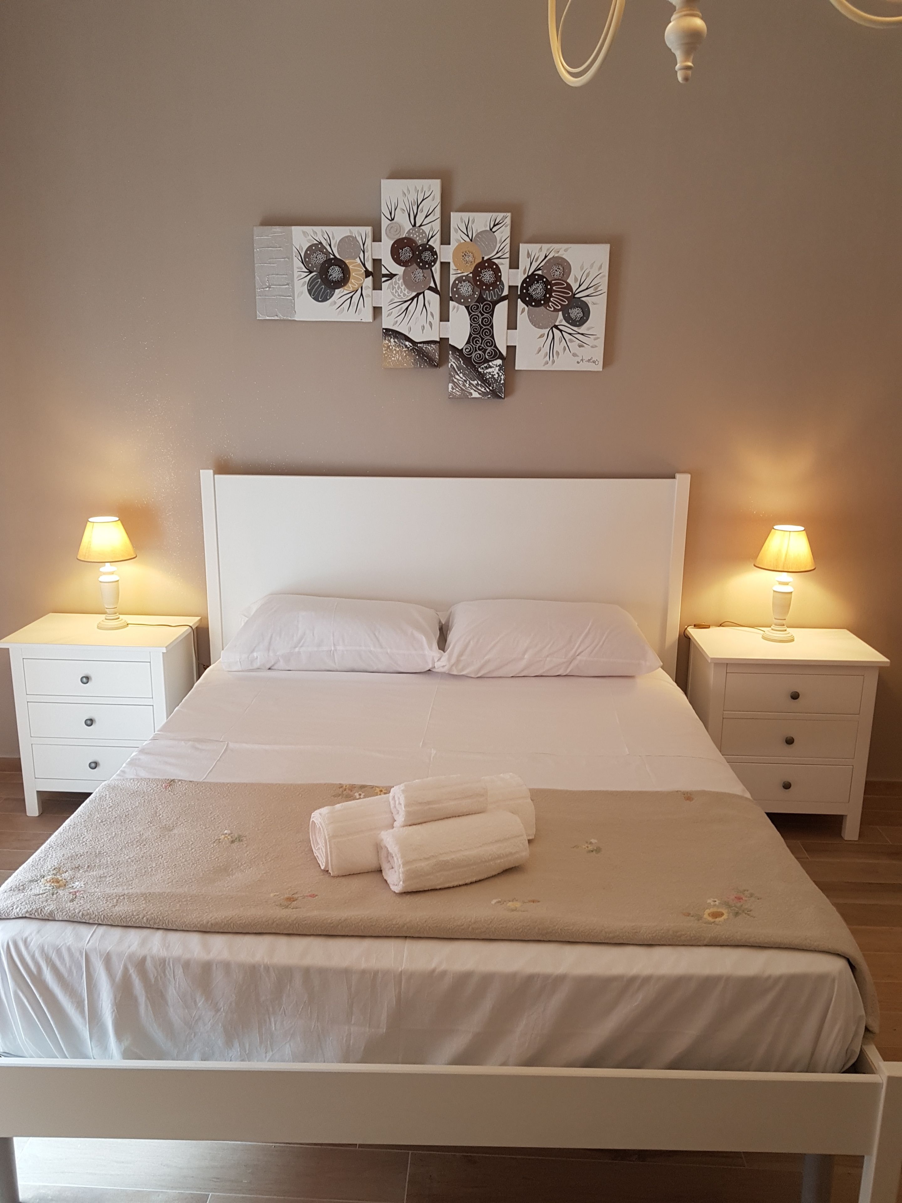 Una cama bien hecha con sábanas blancas, dos mesitas de noche con lámparas y una obra de arte colgada en la pared sobre la cama.