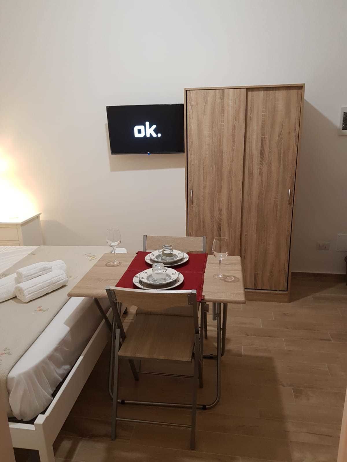 Een kleine, verzorgde kamer met een bed, een gedekte tafel voor twee met borden en glazen, een aan de muur bevestigde televisie en een houten garderobe.