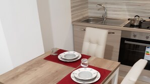 Zona pranzo con tavolo in legno, sedie bianche e sottopiattino rosso. Sullo sfondo, una cucina con lavello, piano cottura e pensili.