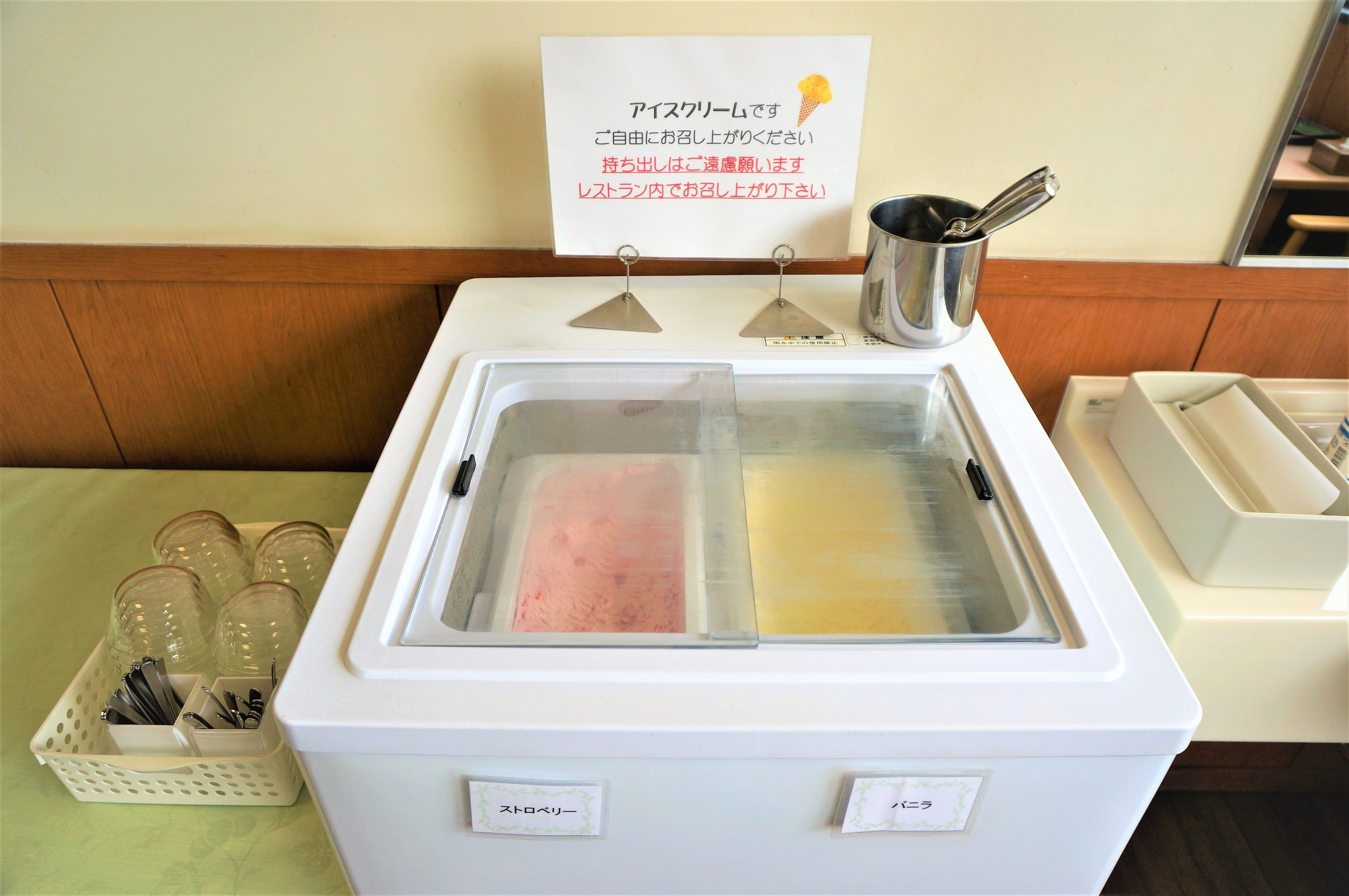 Máquina expendedora de helados de servicio propio, con dos compartimentos, uno rosa y uno amarillo. Se observa un letrero en un idioma extranjero sobre la máquina, y una cesta con utensilios a la izquierda.