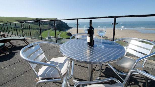 Mesa redonda de vidrio con sillas de metal, una botella de vino y dos copas en un balcón con vistas a una playa y acantilados.