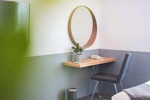 A hotel room with a green wall, a wooden desk with a telephone and a potted plant, a black chair, and a trash bin.