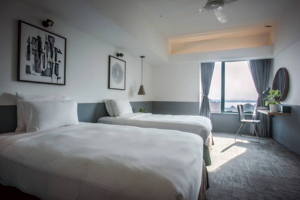 A hotel room with two beds, a desk with a chair, a large window with a view, and artwork on the walls.