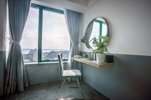 A hotel room with a view of a cityscape through a large window, a desk with a chair, a mirror, and a potted plant.