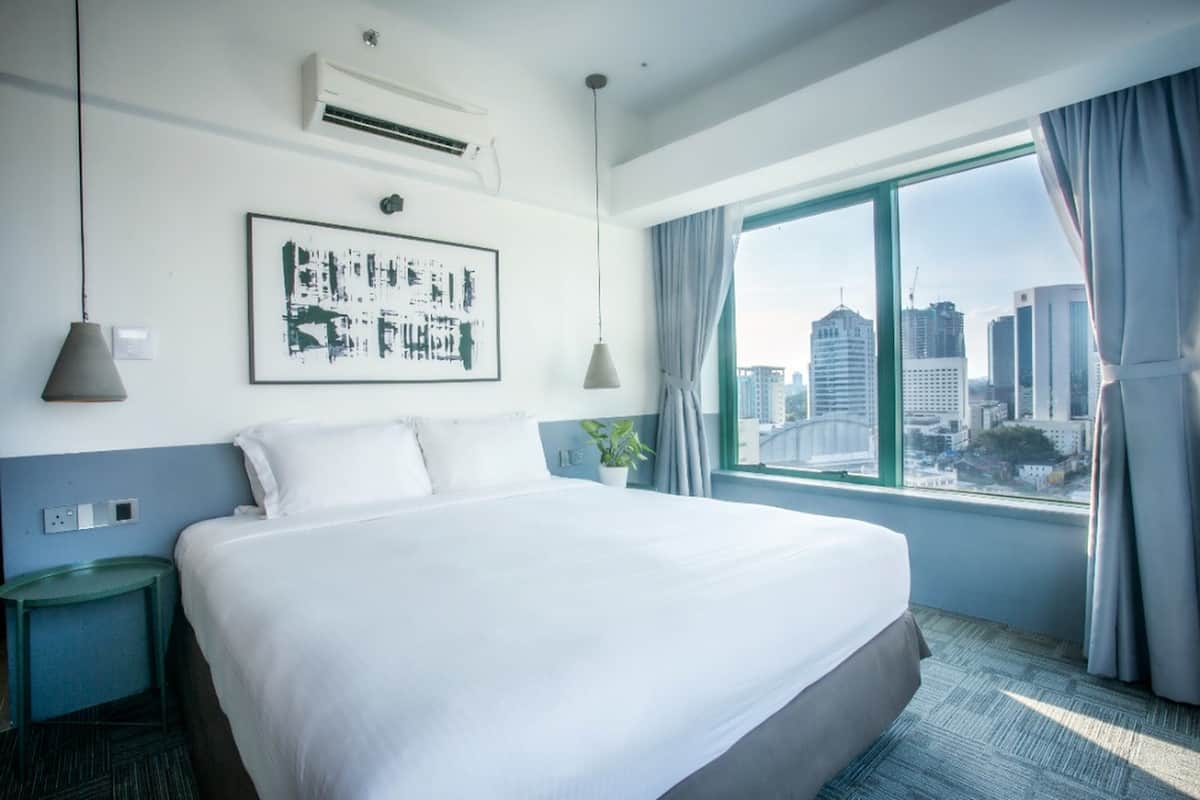 A hotel room with a large bed, a city view, an air conditioner, a framed artwork, and a small table with a plant.