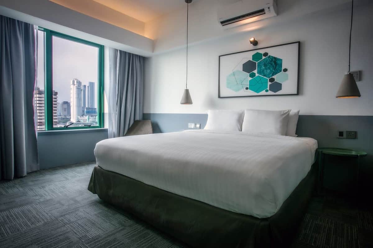 A hotel room with a large bed, a view of the cityscape, a modern art piece on the wall, and a green side table.