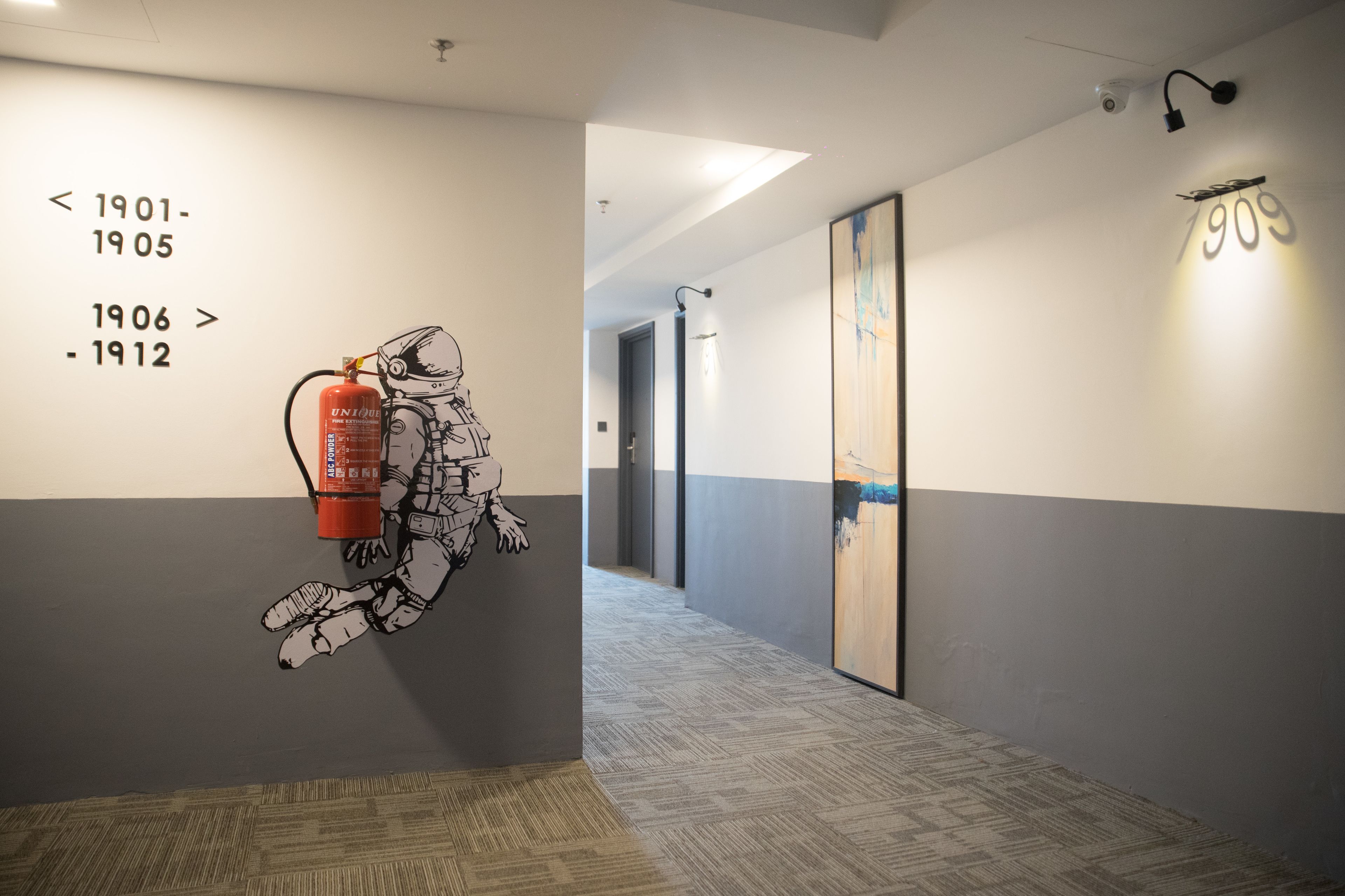 A hallway with a wall-mounted artwork of a firefighter and a fire extinguisher, a corridor with a patterned carpet, and a framed painting on the wall.
