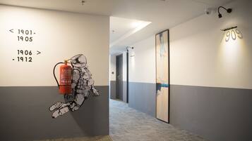 A hallway with a wall-mounted artwork of a firefighter and a fire extinguisher, a corridor with a patterned carpet, and a framed painting on the wall.