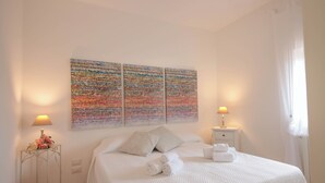 Een net opgemaakt bed met witte lakens en kussens, een nachtkastje met een lamp, en een kleurrijk abstract schilderij erboven.