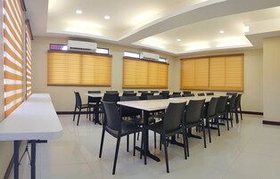 Un salón de conferencias con varias mesas y sillas, unidades de aire acondicionado y grandes ventanales con persianas.
