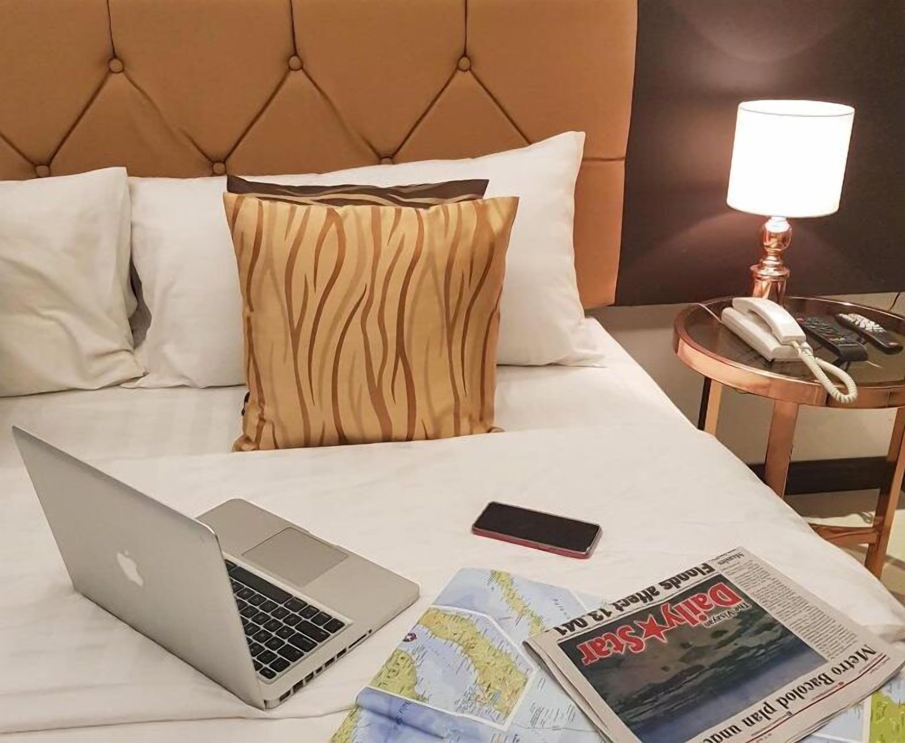 A hotel room with a bed, a laptop, a mobile phone, a newspaper, and a bedside table with a lamp and a telephone.