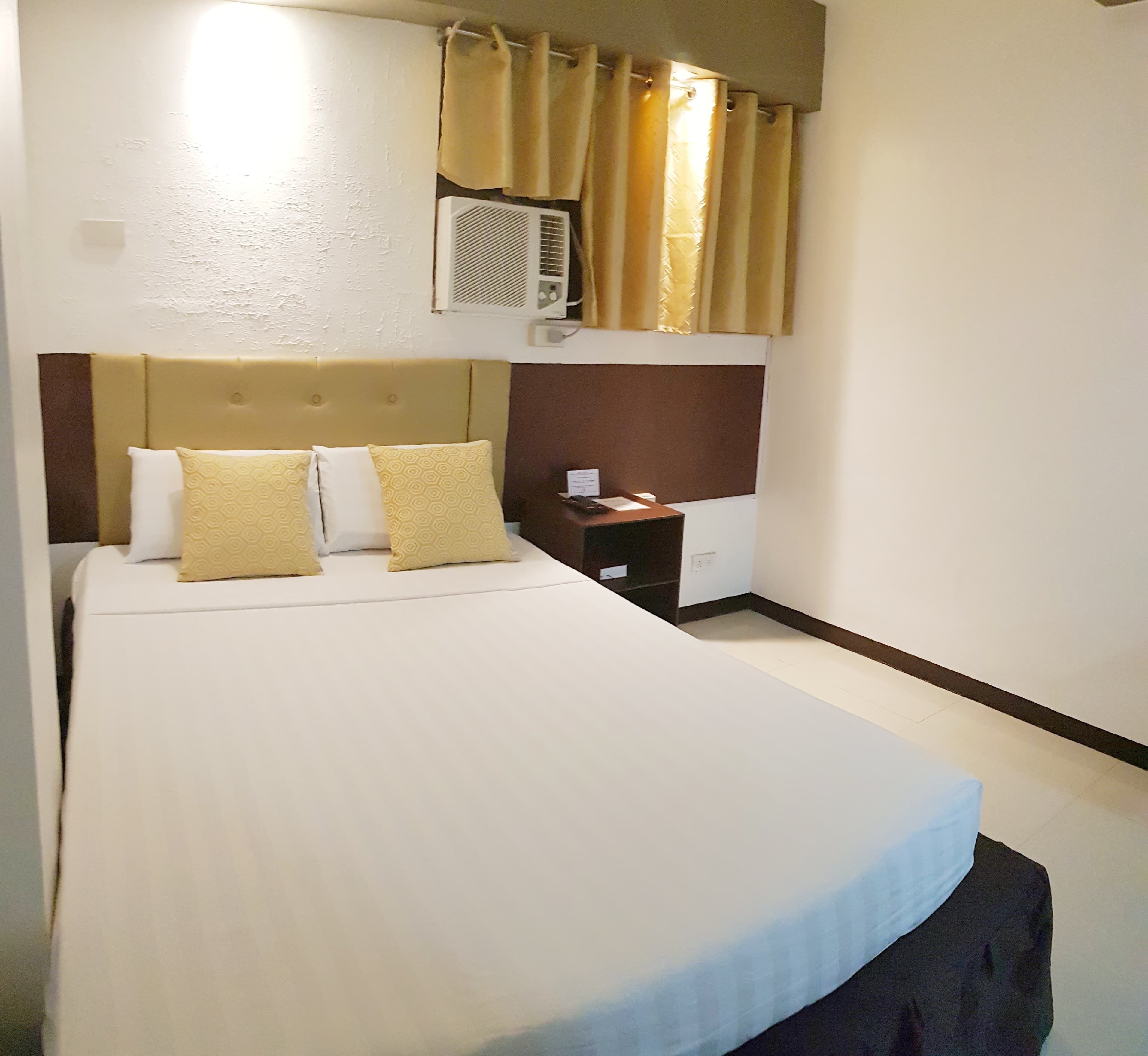 A hotel room with a large bed, a bedside table, a nightstand, a wall-mounted air conditioner, and a window with curtains.