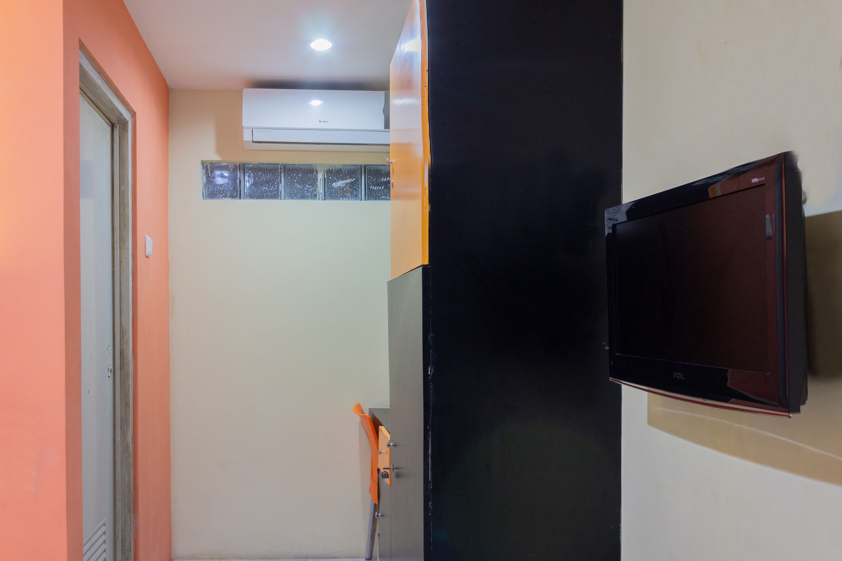 Una habitación con un televisor montado en la pared, un equipo de aire acondicionado sobre una ventana y una puerta a la izquierda.