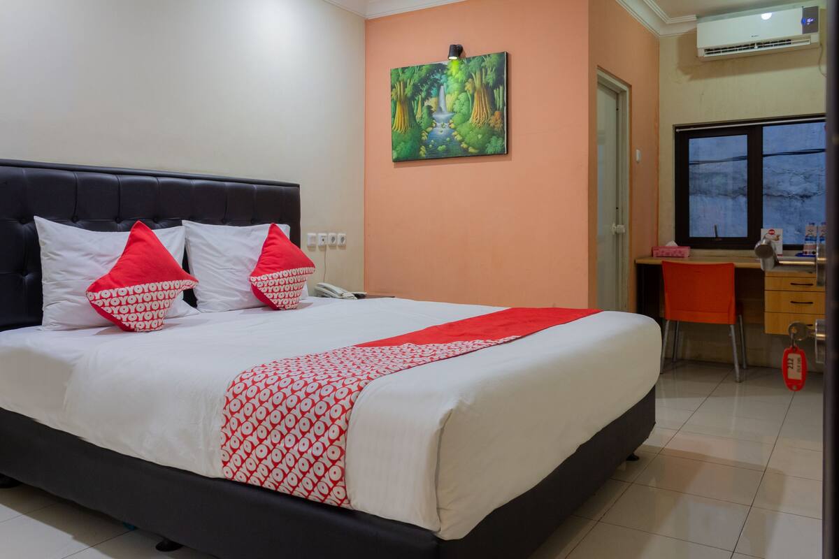 A neatly made bed with white and red bedding, a painting of a forest scene on the wall, and a desk with a chair in the corner.