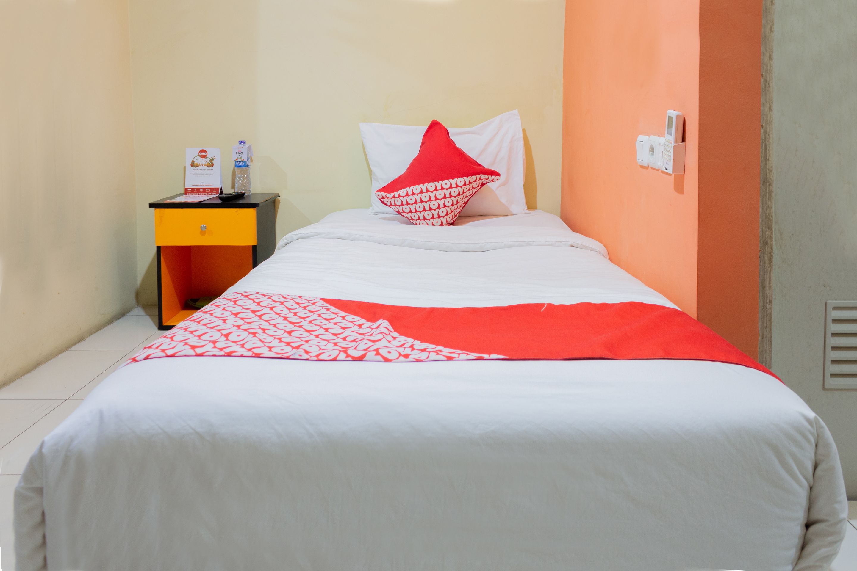 A hotel room with a bed, a nightstand with a phone and a small table, and a wall-mounted light switch.