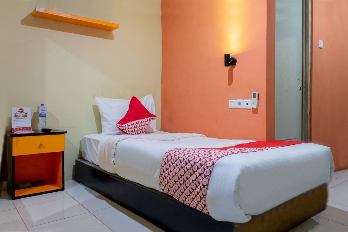 A hotel room with a bed, a nightstand, a shelf, and a wall-mounted light.