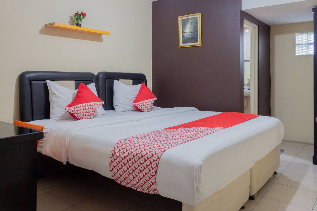 A neatly made bed with a red and white patterned bedspread, two pillows with red and white designs, and a small shelf with flowers above it.