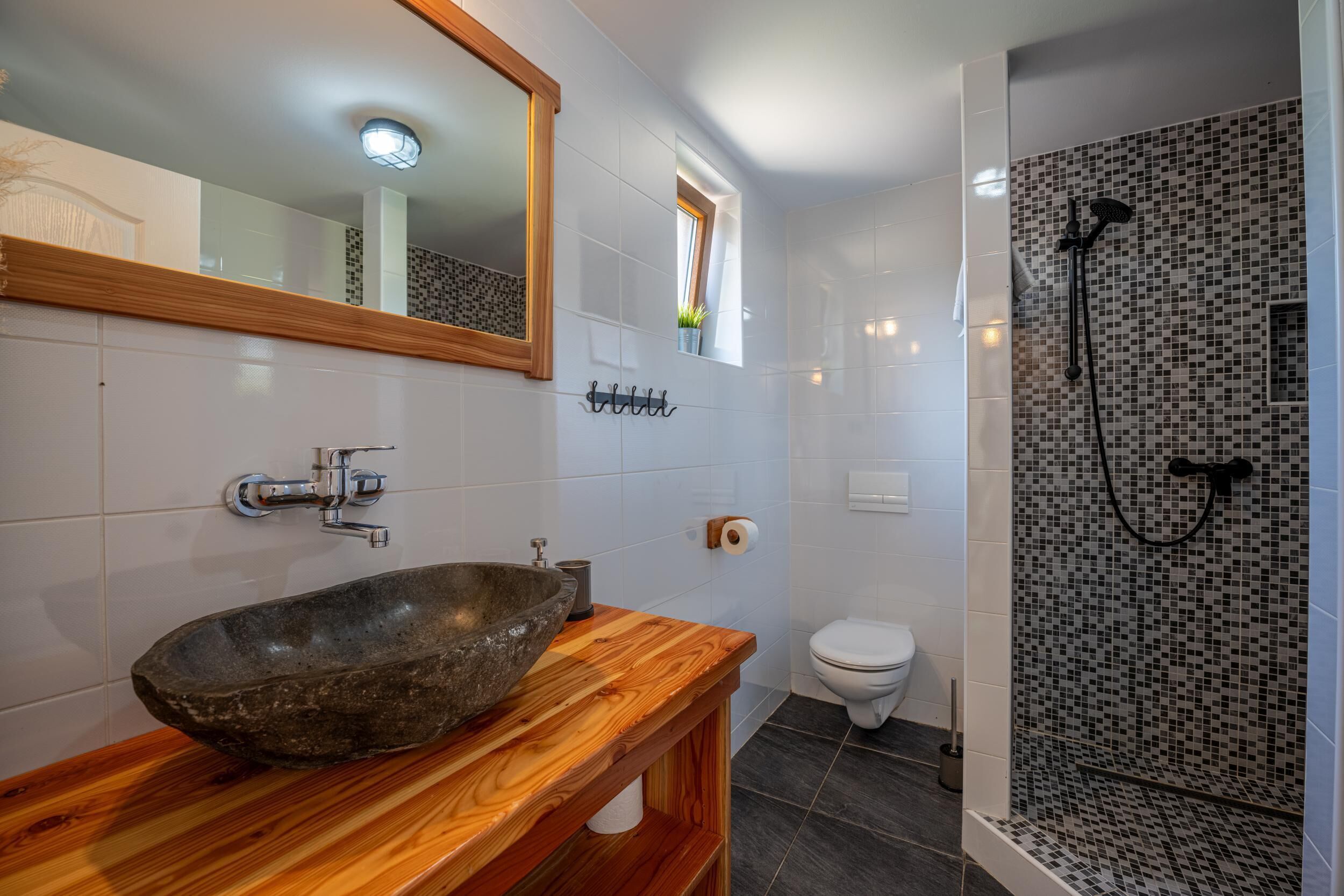 A bathroom with a stone basin, a wooden vanity, a toilet, and a shower area with mosaic tiles.