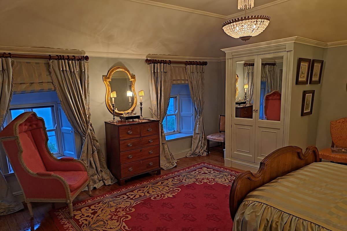A bedroom with a large bed, a vanity with a mirror, a red armchair, a wooden dresser, and a chandelier.