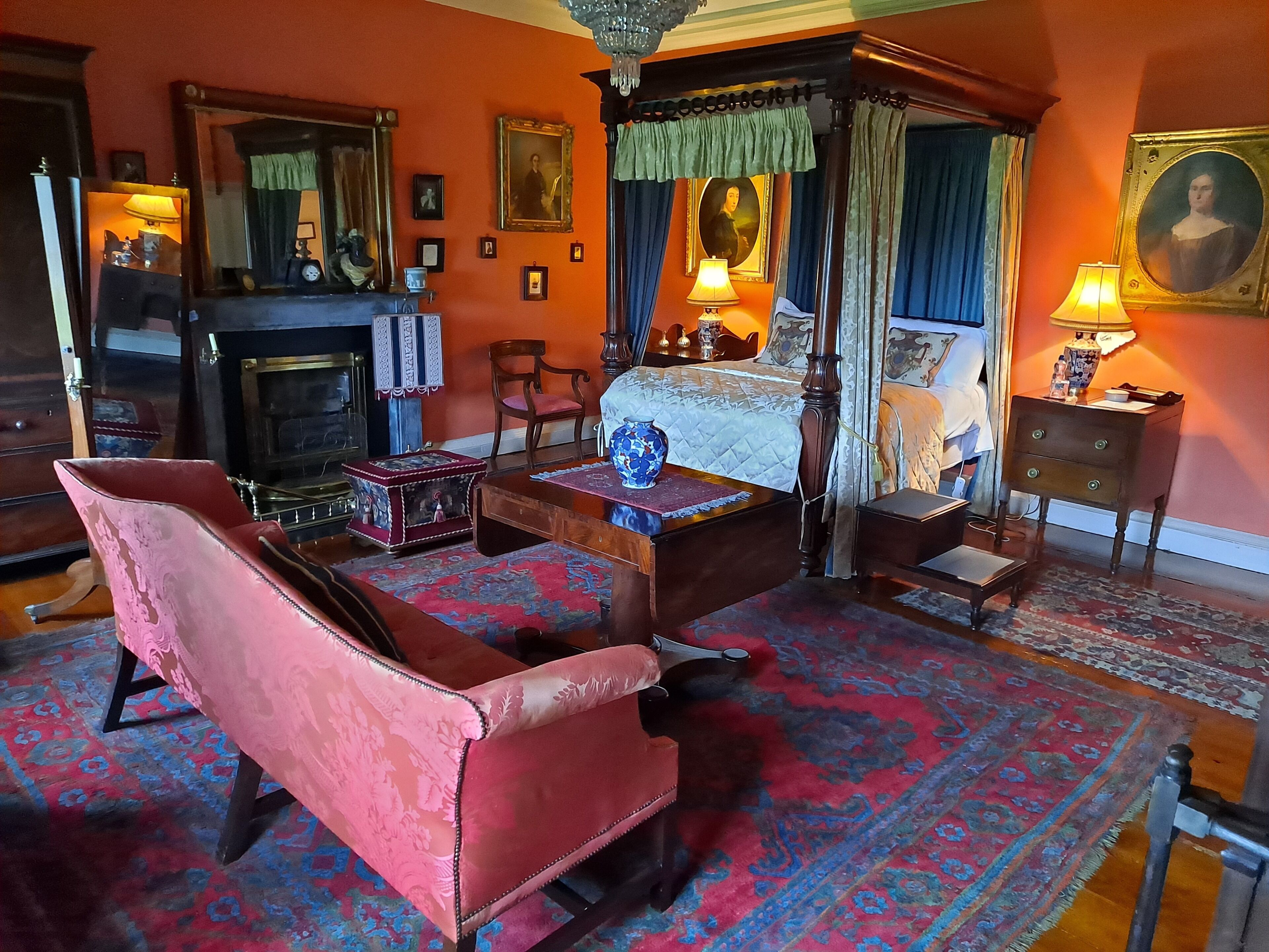 A traditional bedroom with a four-poster bed, a sitting area with a red armchair, a wooden chest, and a fireplace.