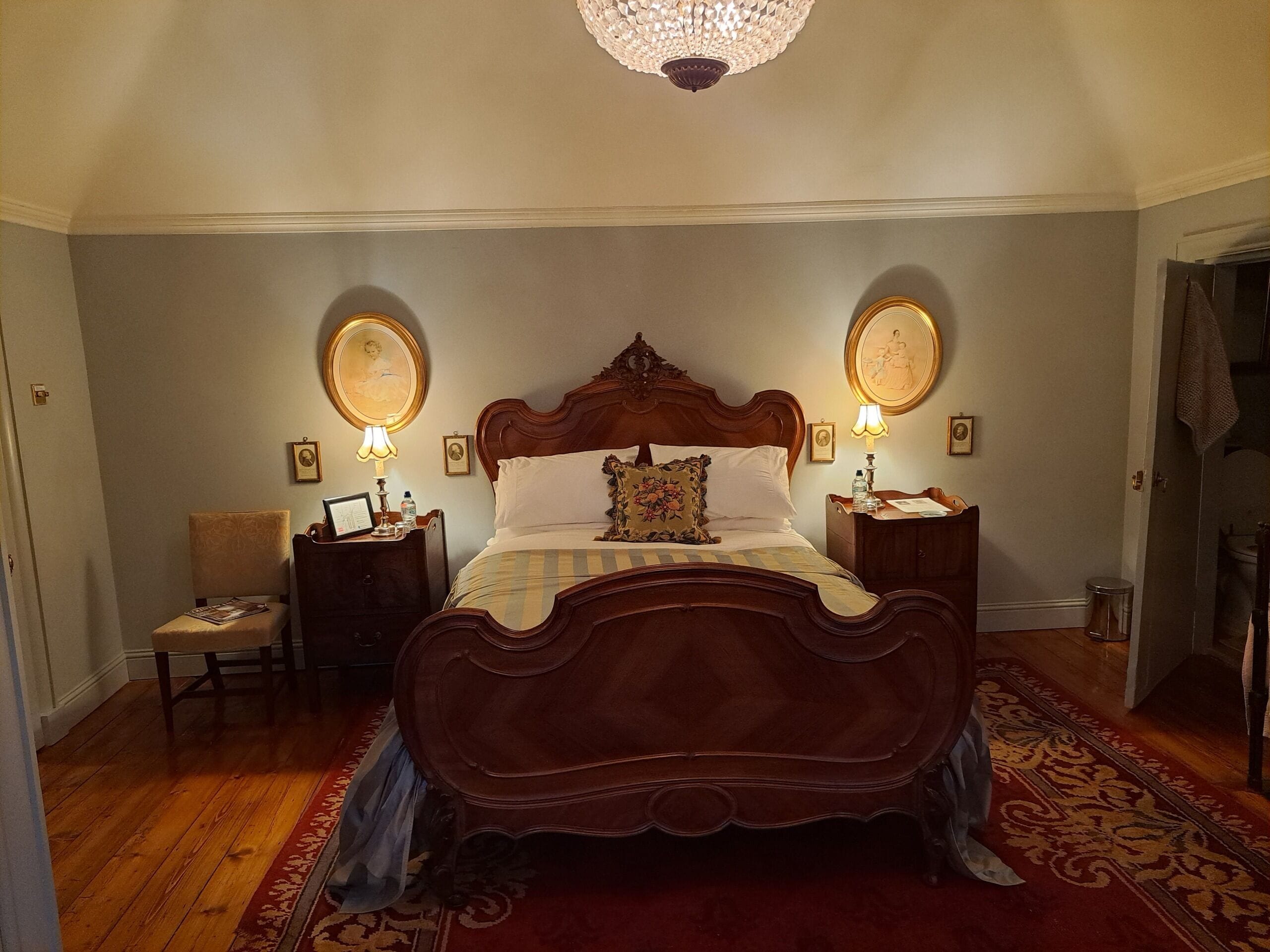 A bedroom with a large wooden bed, two bedside tables with lamps, a chair, and a chandelier.
