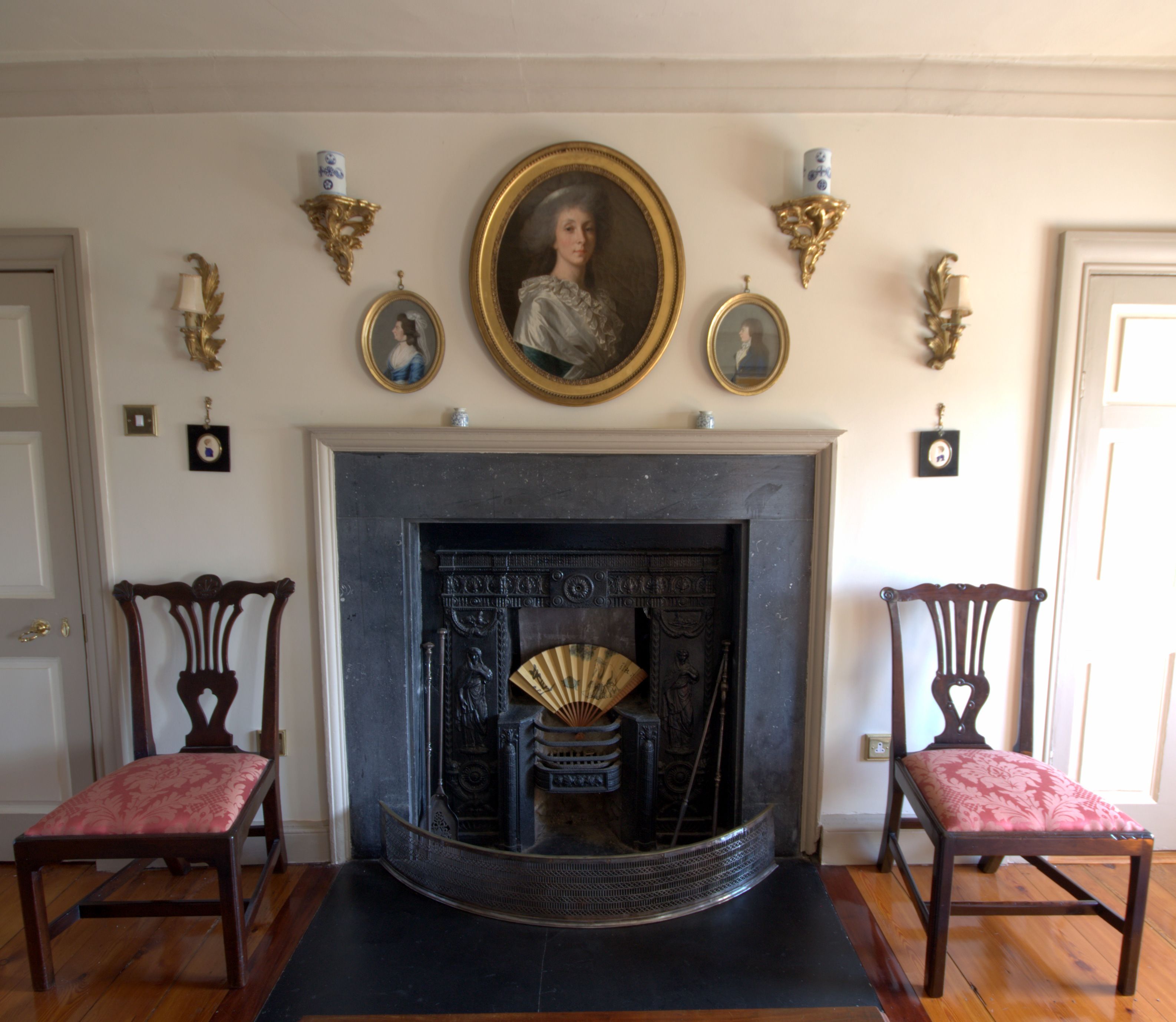 A room with a fireplace, two chairs, and a portrait above the fireplace.