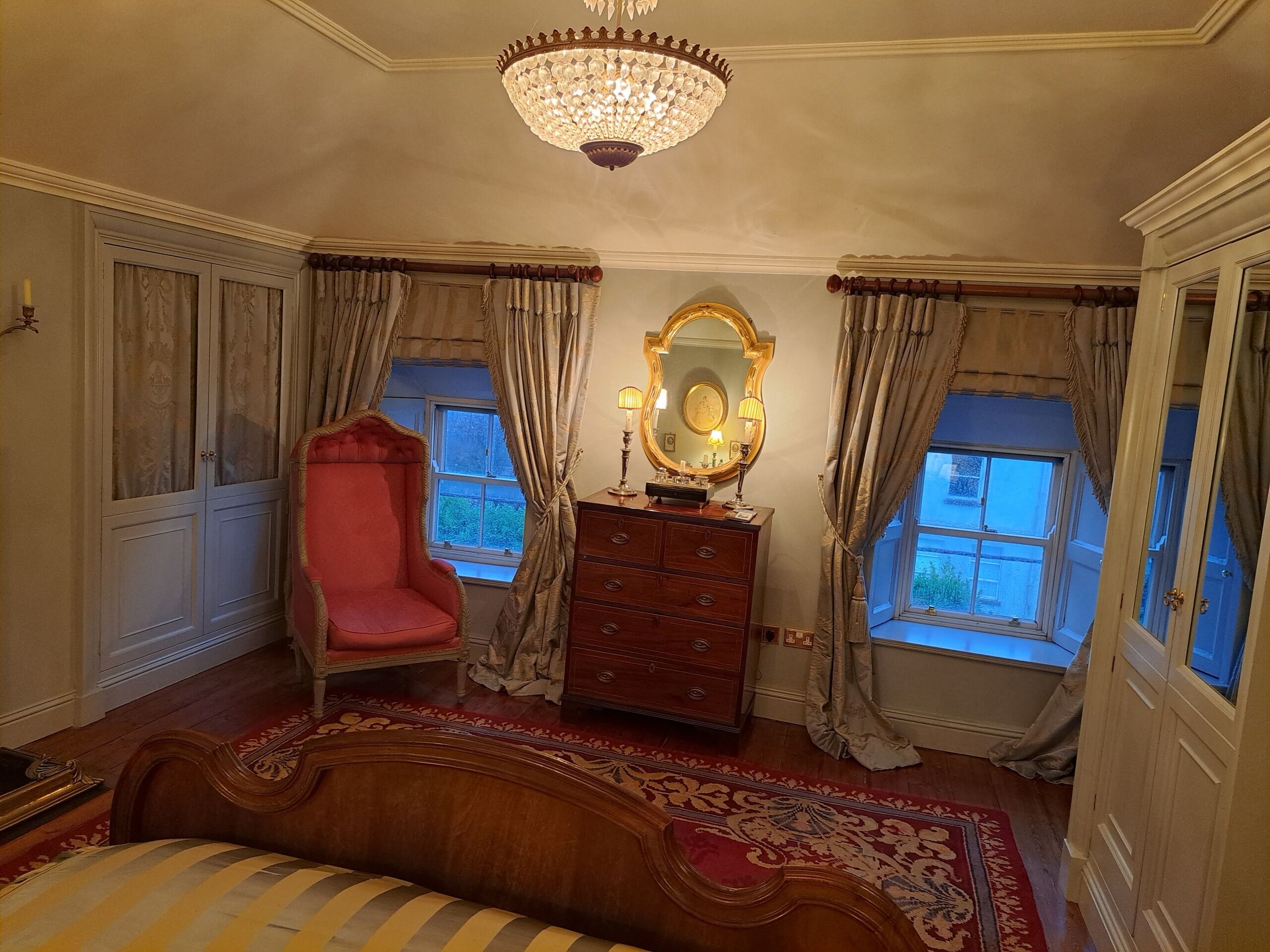 A bedroom with a large mirror, a red chair, a dresser, and a chandelier.