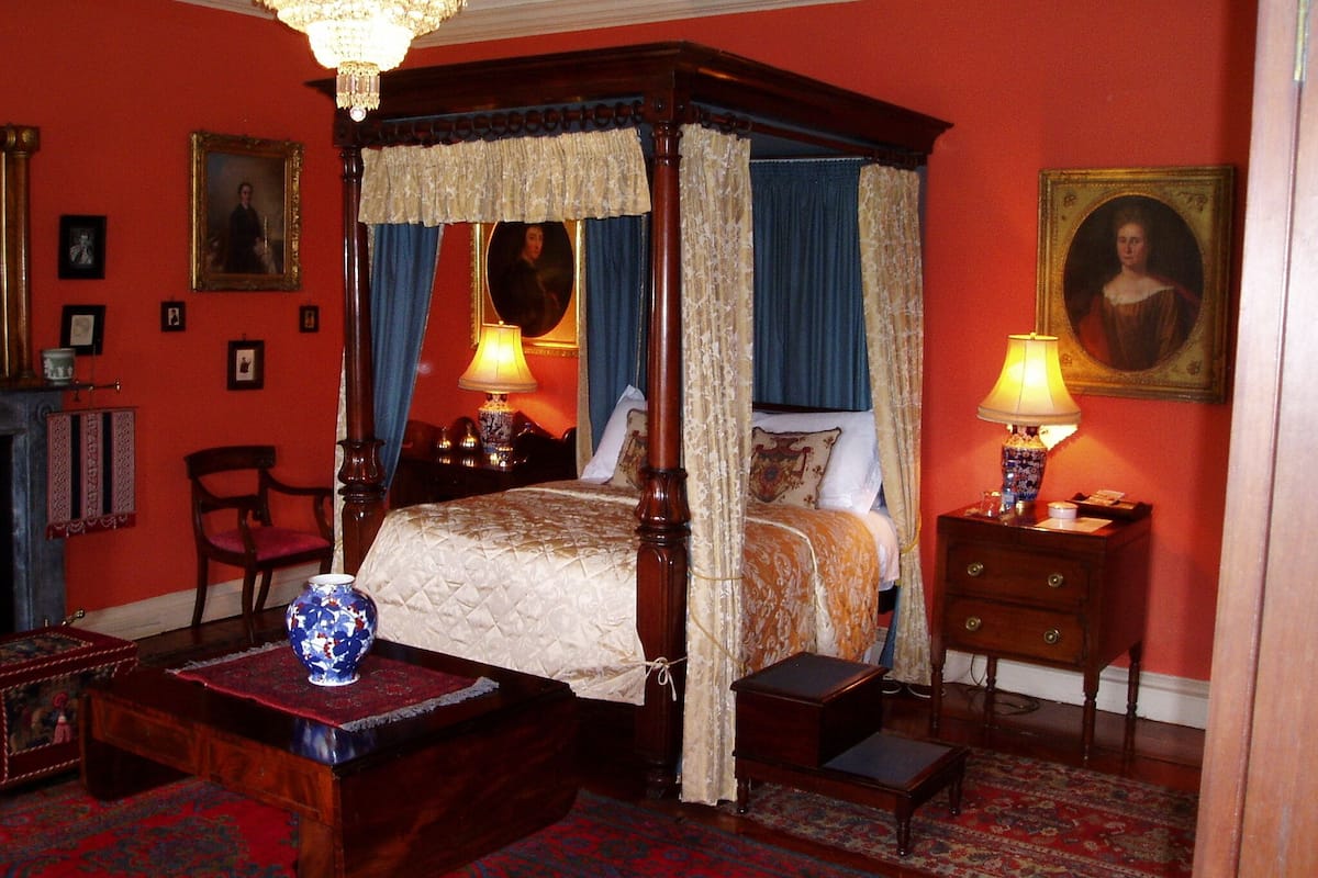 A four-poster bed with a canopy, a wooden nightstand with a lamp, a chair, and a small table with a vase.