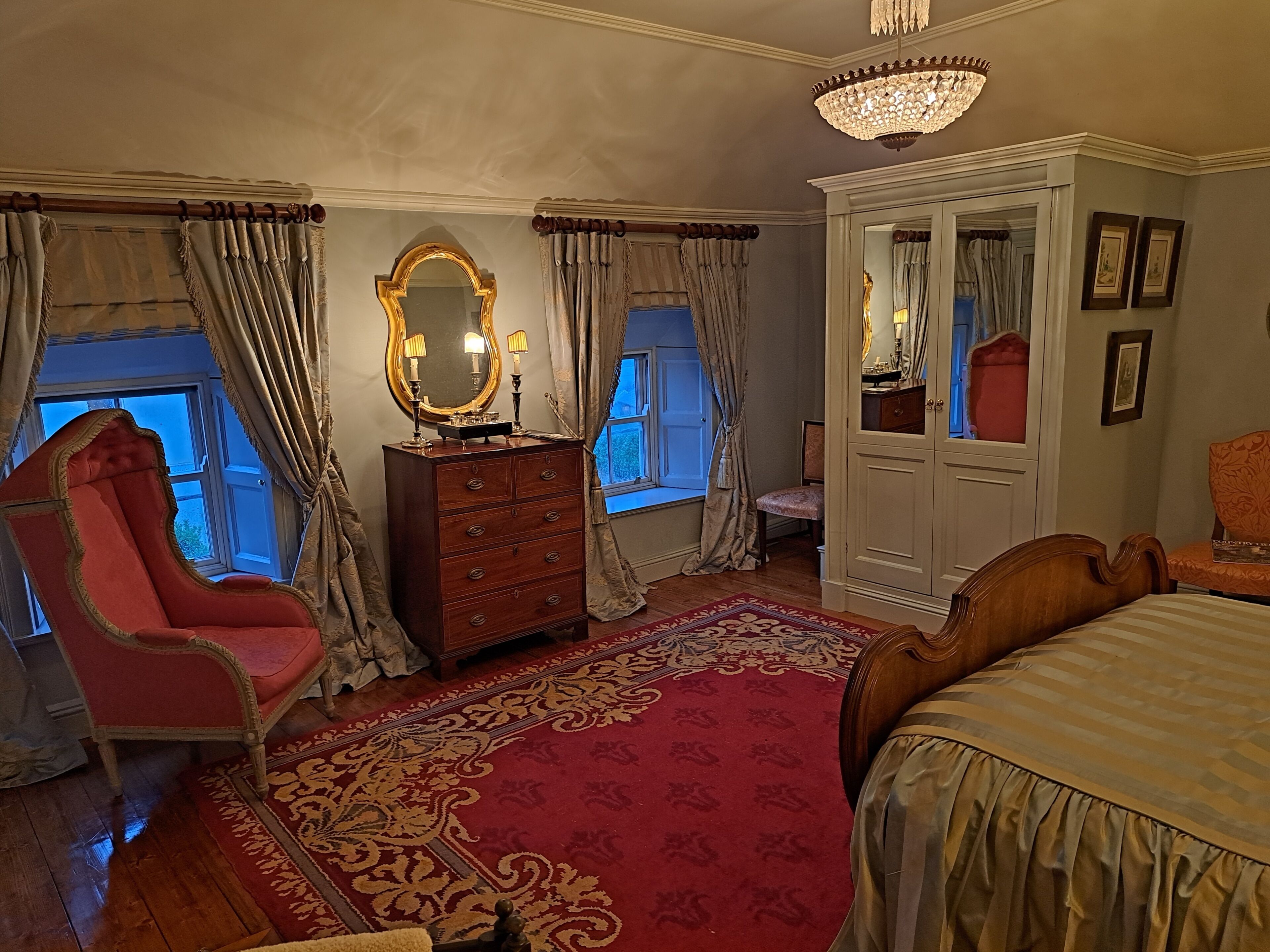 Un dormitorio con una cama grande, una tocador con espejo, un sillón rojo, un aparador de madera y una alfombra roja estampada.