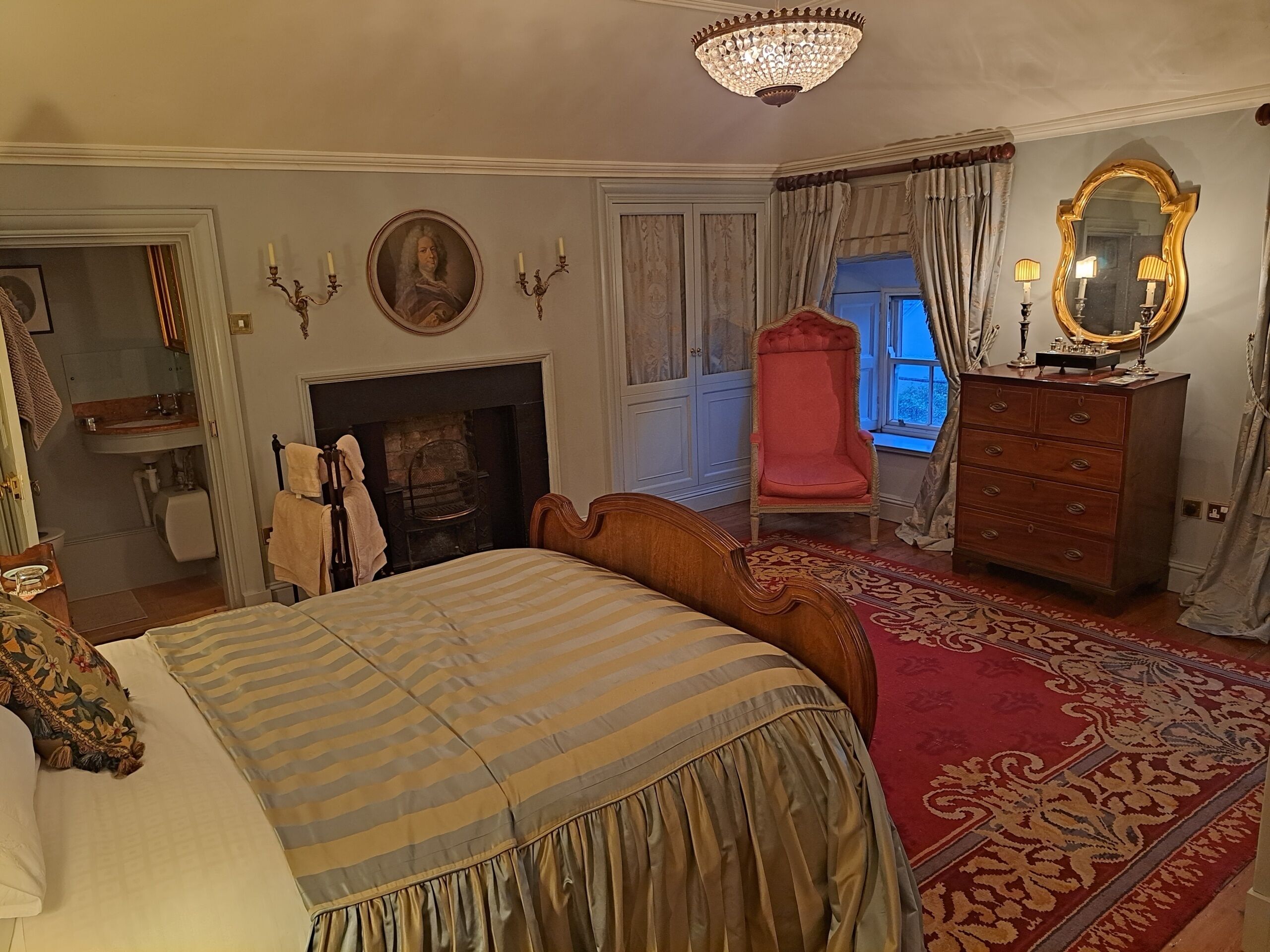 A traditional bedroom with a bed, a fireplace, a red armchair, a dresser, and a mirror.