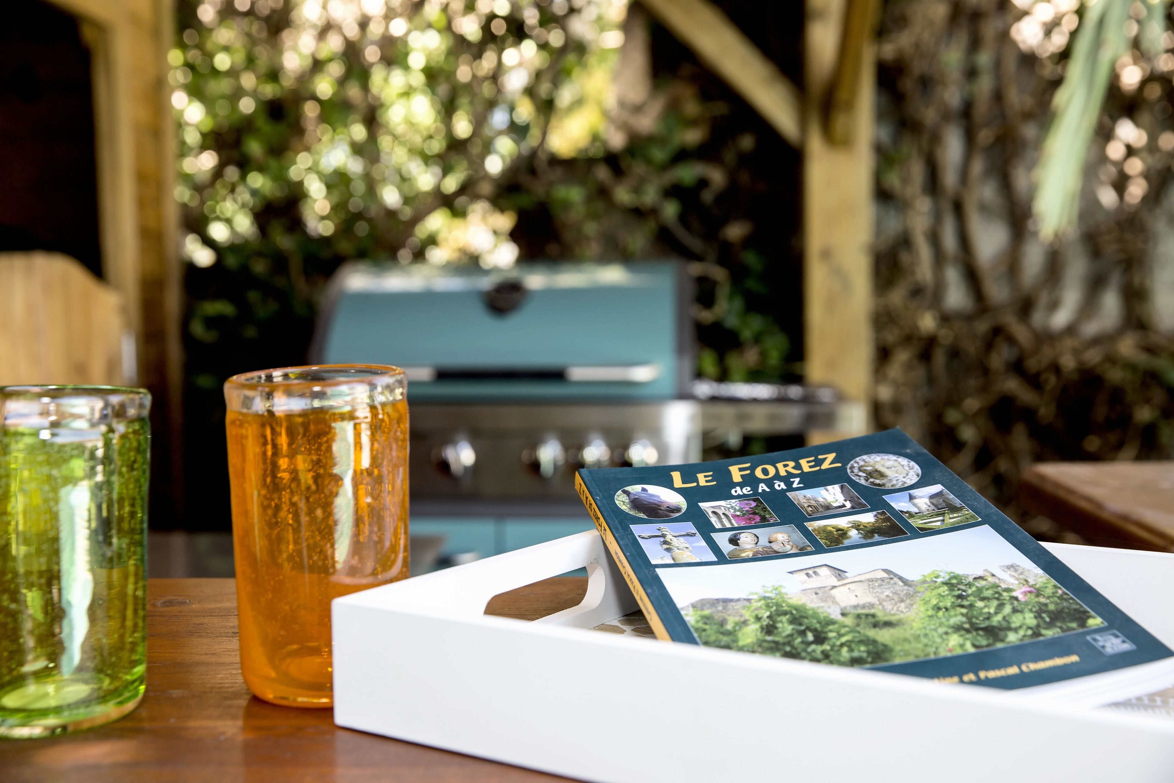 A table with a book titled "Le Forez de A", two glasses of beverages, and a grill in the background.