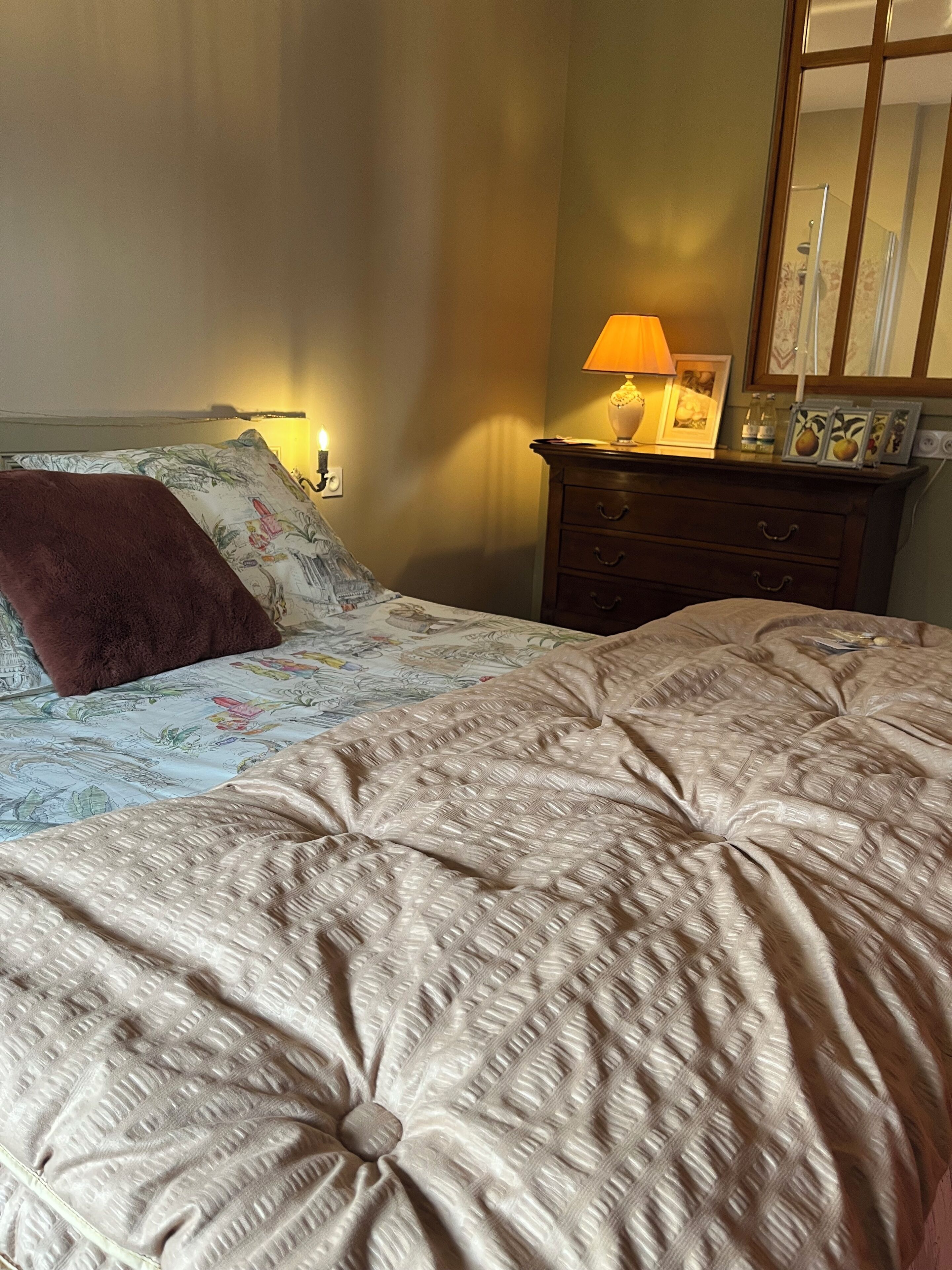 Un lit bien fait, recouvert d’une couvre-lit à motifs et agrémenté d’un coussin décoratif. On aperçoit une table de chevet avec une lampe et un tableau encadré. Un miroir est visible sur le mur.