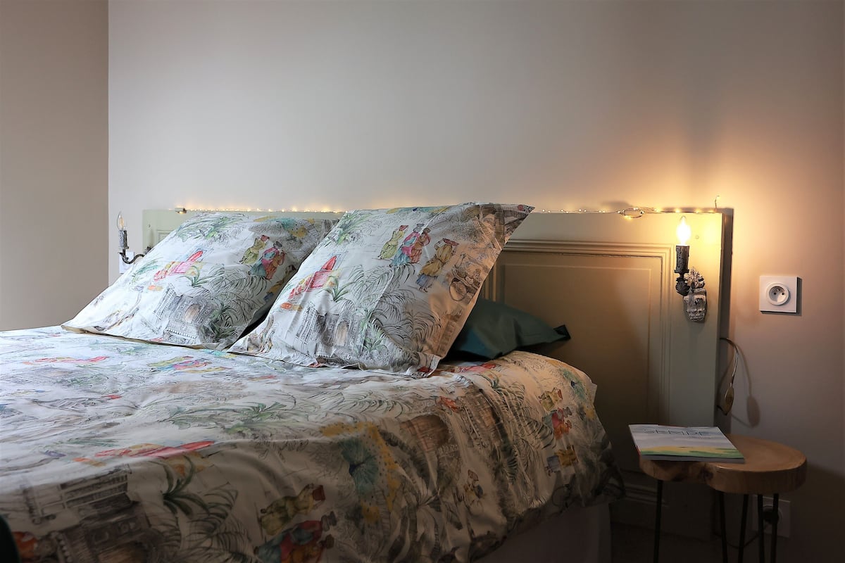 Una cama con una colcha y almohadas de estampado floral, una mesita de noche con un libro, y una lámpara de pared.