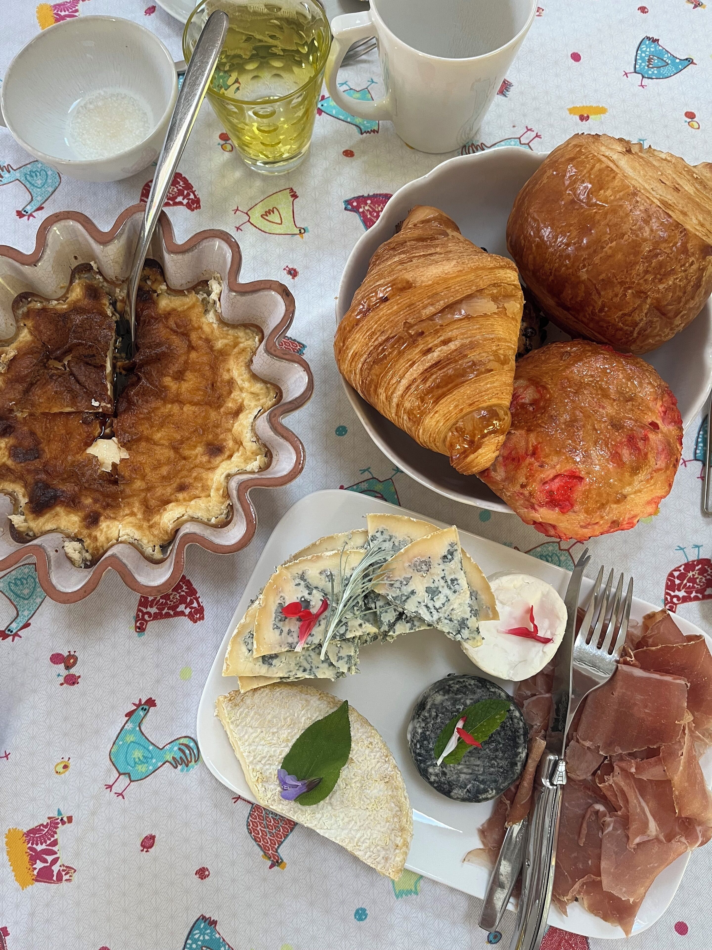 Ein Frühstückstisch mit Quiche, Croissants, Käse und Schinken, gedeckt mit einer bunten Tischdecke.