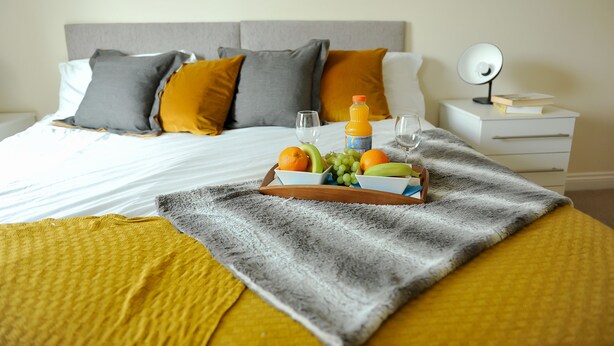 A neatly made bed with a tray of fruit and juice, a bedside table with a lamp, and a bedside book. - Select Serviced Accommodation - Hunters Wharf (Reading)