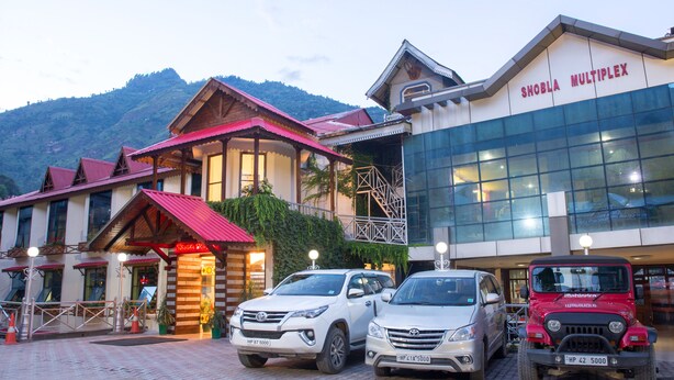 A hotel with a traditional wooden facade, a modern glass building, and parked cars in front. - Hotel Shobla Royale (Kullu)