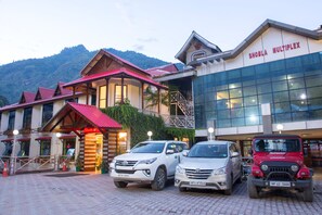 A hotel with a traditional wooden facade, a modern glass building, and parked cars in front. - Hotel Shobla Royale (Kullu)
