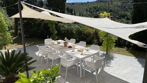 An outdoor dining area with a table set for four, surrounded by white chairs, under a large shade sail.